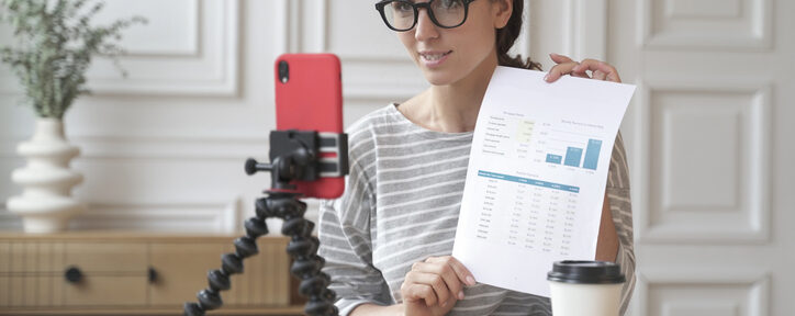 Female accounting influencer recording video content while holding financial charts and graphs, with smartphone on tripod and laptop on desk in home office setting