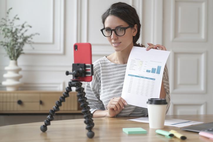 Female accounting influencer recording video content while holding financial charts and graphs, with smartphone on tripod and laptop on desk in home office setting