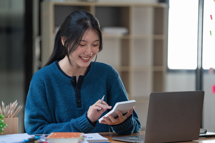 Female accounting student smiling while reviewing notes on tablet, sitting at desk with laptop and office supplies in modern study space